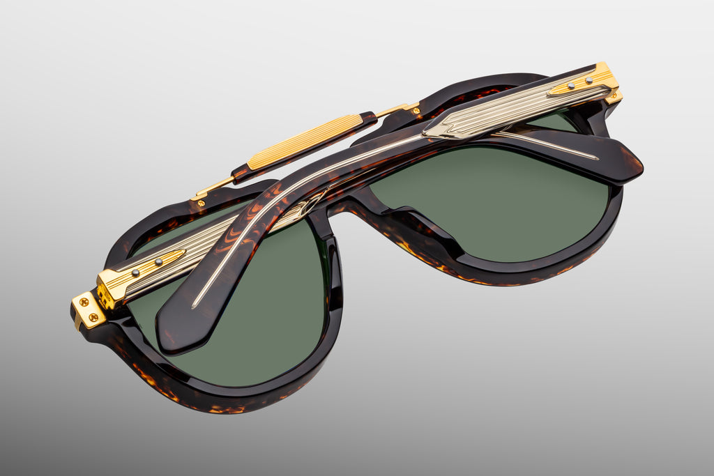 Dark brown acetate frame in tortoiseshell pattern with gold hardware across temples and brow bar, and green sunglass lenses laying facedown on a grey background