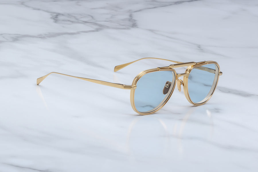 3/4 angle view of a gold metal aviator frame with light blue lenses and a gold brow bar, on top of a white marble background.