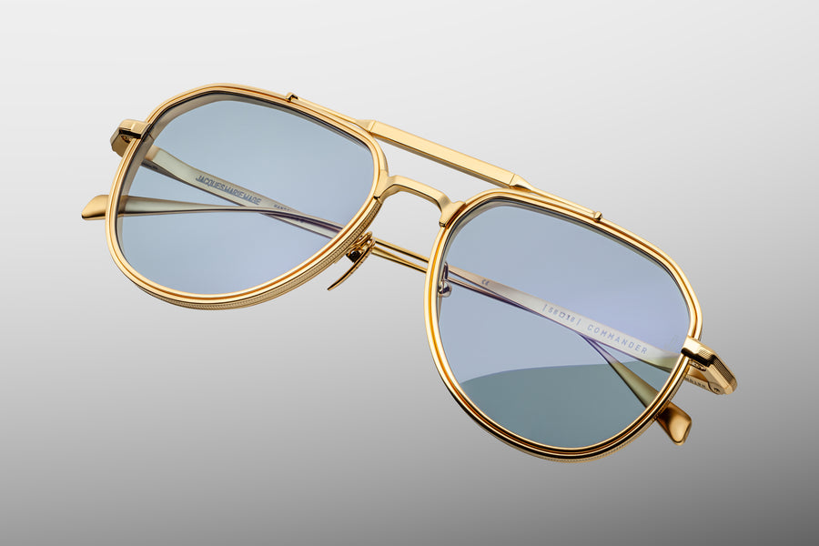 Gold aviator frame with a golden brow bar and light blue lenses, floating at a diagonal on top of a grey background. 