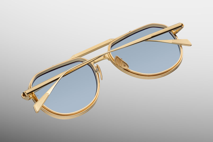 Back view of a gold aviator frame with blue lenses, a gold brow bar, and think temples folded over one another, floating at a diagonal on top of a grey background.