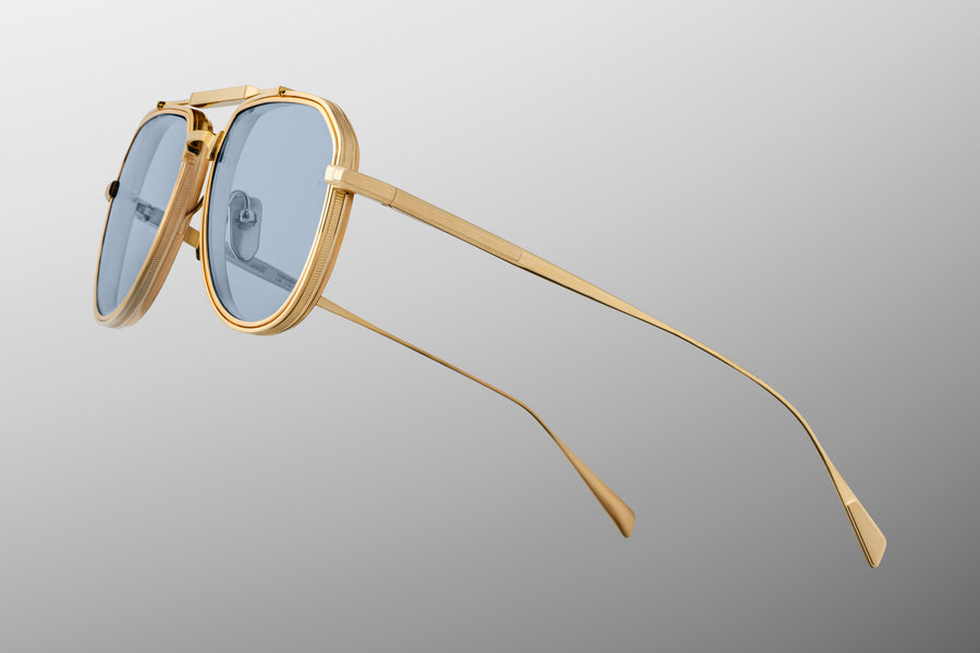 Side profile of a gold metal aviator frame with blue lenses and thing temples, floating at a diagonal on top of a grey background.