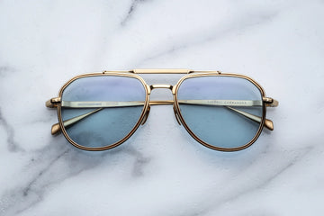 Gold metal aviator frame with light blue lenses, on top of a white marble background.