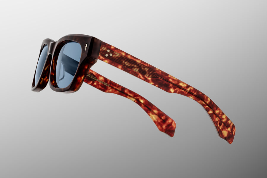 Side view of reddish brown tortoiseshell acetate sunglasses with blue lenses floating against a grey backdrop