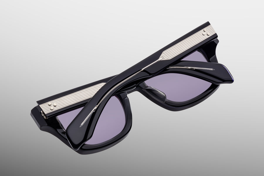 Dark blue sunglasses with purple lenses and bold silver exposed wirecore, lens down against a grey backdrop 