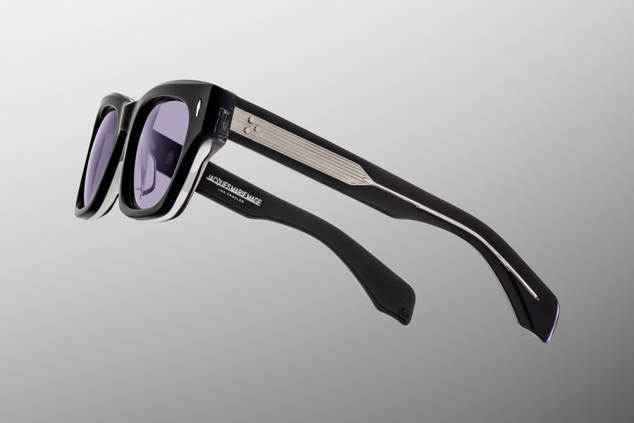 Side view of dark blue acetate sunglasses with purple lenses and exposed silver hardware, floating against a grey backdrop