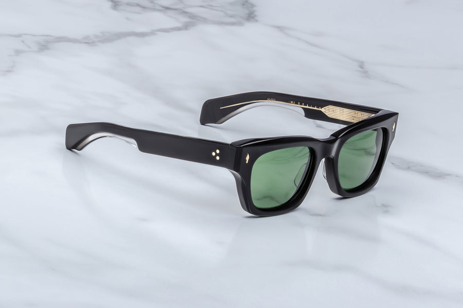 Side profile of black acetate sunglasses with green lenses and visible gold wirecore, across a marble background