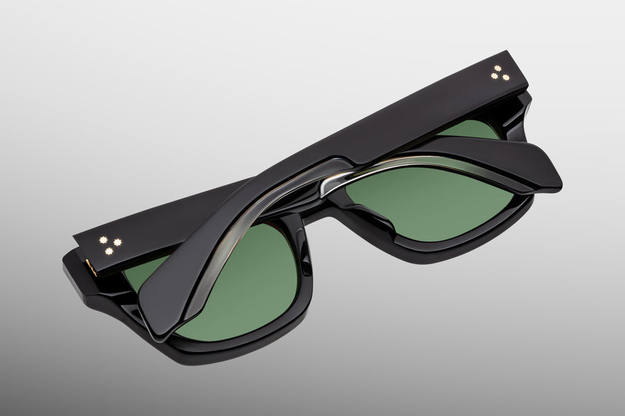 Black sunglasses with green lenses and crystal highlights, lens down against a grey backdrop 
