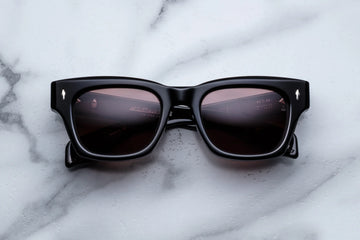 Thick black acetate frame with deep brown sunglass lenses on a white marble background