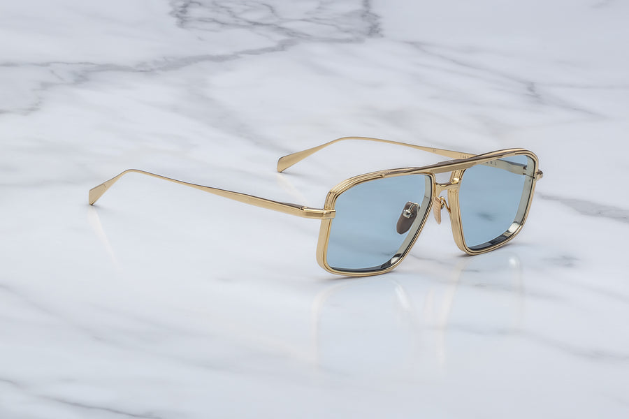 3/4 view of a gold square-aviator frame, with light blue lenses and thin temples, on top of a white marble background.