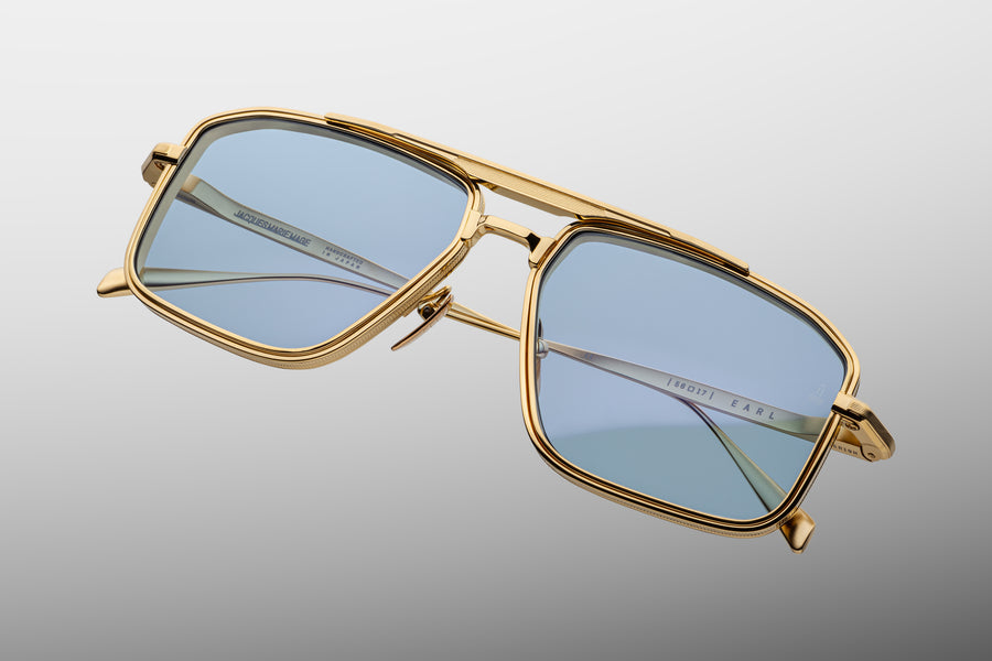 Gold square-shaped aviator frame with light blue lenses and a golden brow bar, at a diagonal floating across a grey background.