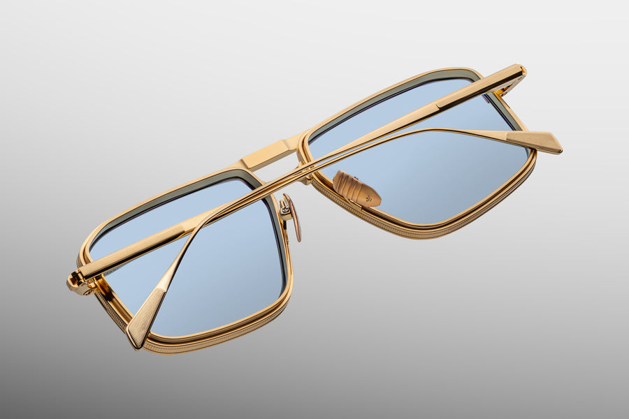 Back view of a gold square-aviator frame, with blue lenses and a prominent brow bar, with thin temples folded over one another across the frame.