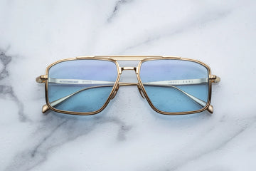 Gold square-shaped aviator frame with light blue lenses and a golden brow bar, on top of a white marble background.