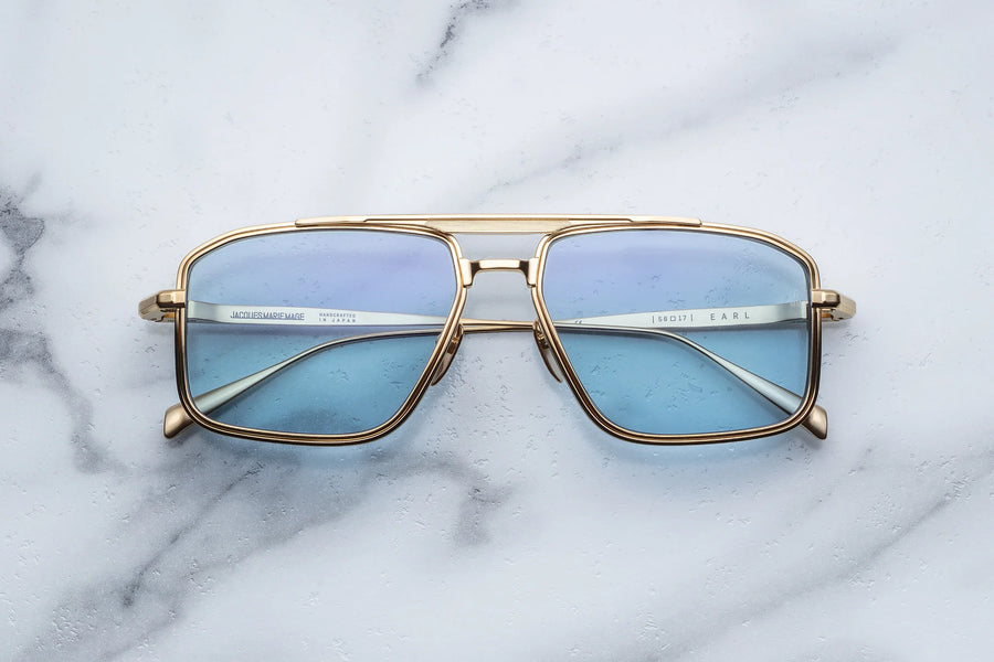 Gold square-shaped aviator frame with light blue lenses and a golden brow bar, on top of a white marble background.
