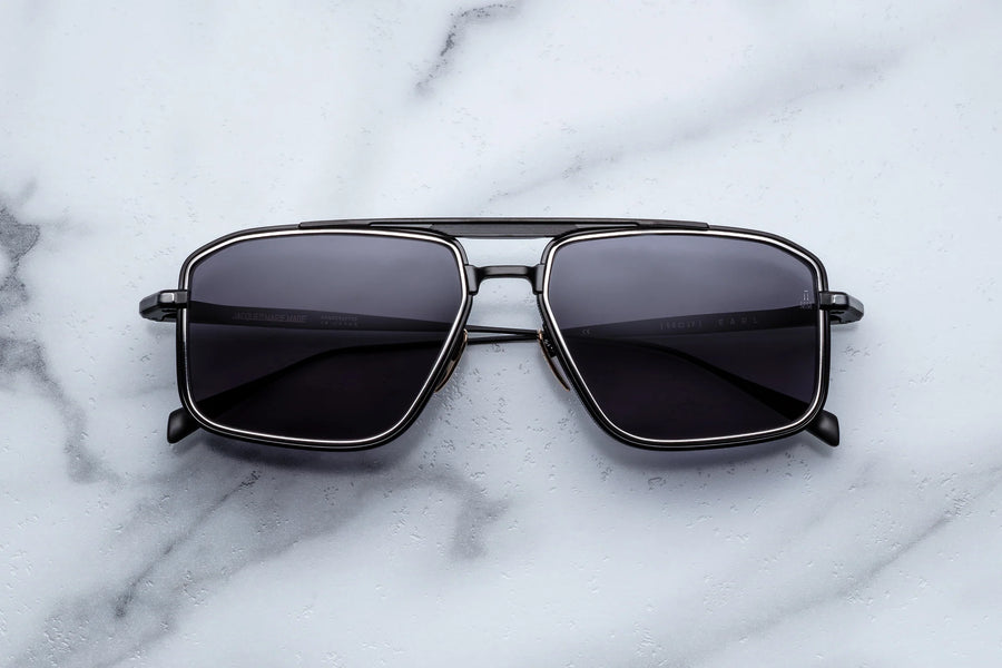 Black metal sunglasses with silver accents in a square aviator shape, laying flat across a white marble background.
