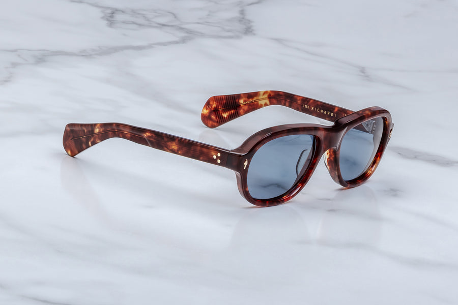 3/4 angle of round aviator style sunglass frames, in a brown tortoiseshell acetate and blue lenses, with light gold hardware, on a white marble background.