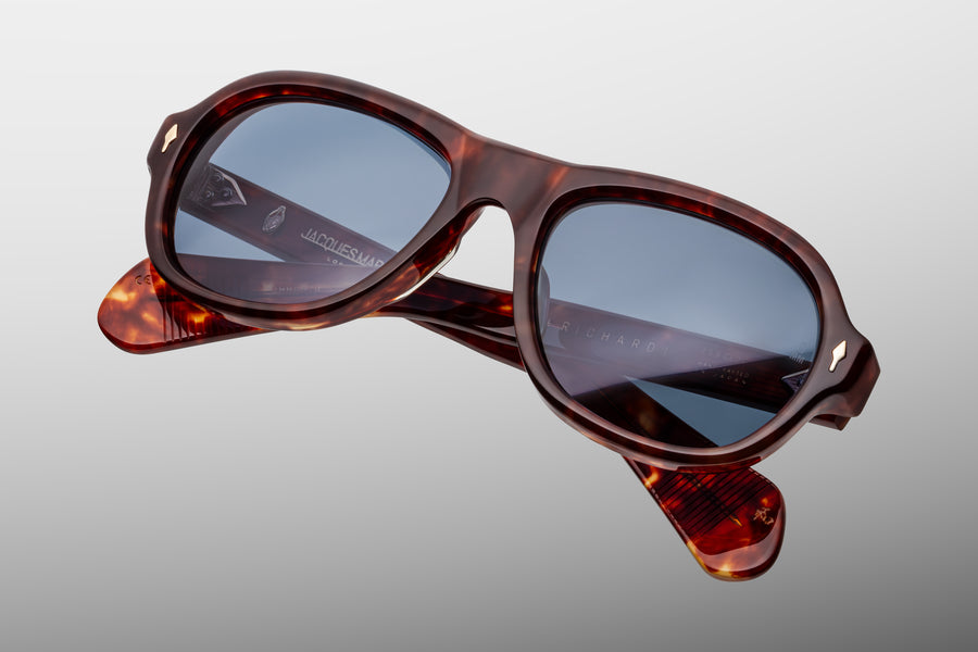 Round aviator style sunglass frame floating against a grey background at a diagonal, frame is in a brown tortoiseshell acetate and has blue lenses.