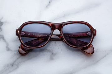 Round aviator style sunglass frame, in a brown tortoiseshell acetate and blue lenses, on a white marble background.