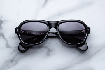 Round aviator style sunglass frame, in black acetate and purple gradient lenses, on a white marble background.
