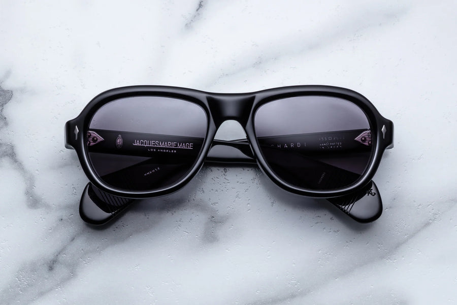 Round aviator style sunglass frame, in black acetate and purple gradient lenses, on a white marble background.