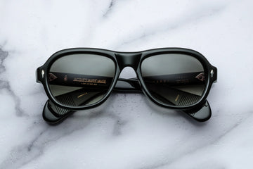 Round aviator style sunglass frame, in green toned black acetate and green gradient lenses, on a white marble background.