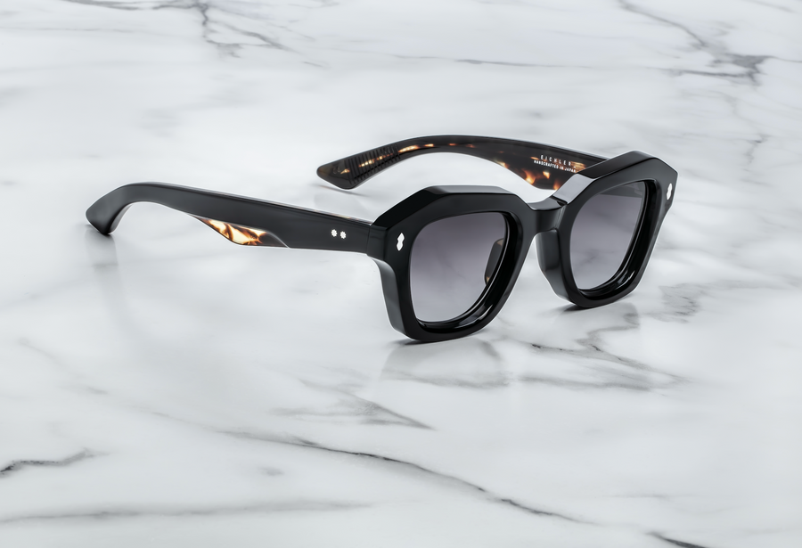 Black sunglasses with silver hardware and tortoiseshell accents on the temples, at a 3/4 angle on top of a white marble background.