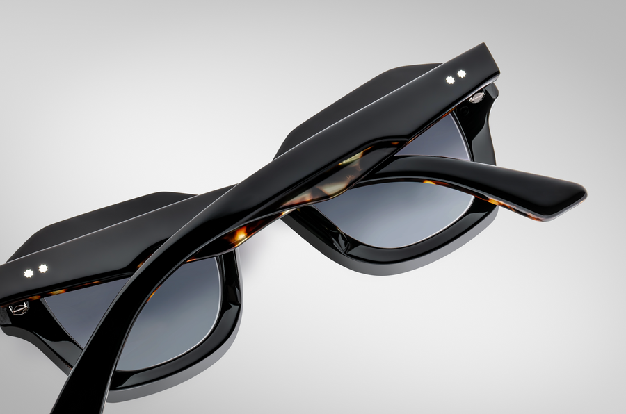 Black sunglasses with tortoiseshell accents on the temples, folded over and pictured laying lens side down on a grey background.