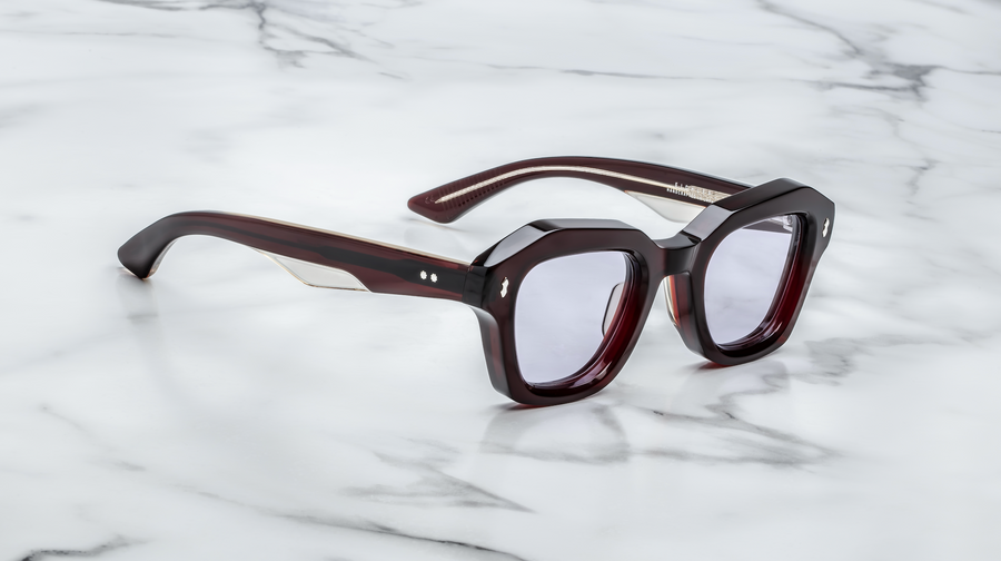 Dark red sunglasses with light purple lenses, at a 3/4 angle on top of a white marble background.