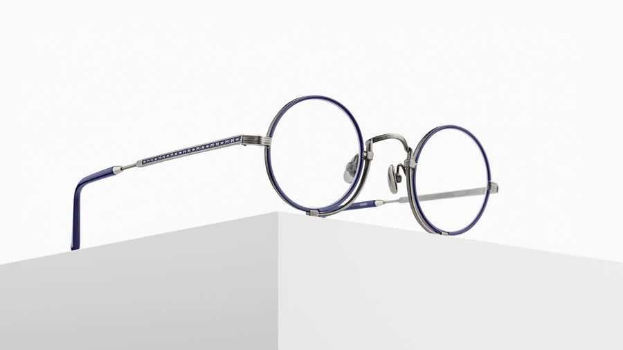 Round eyeglasses with navy blue frames and silver accents on a pedestal against a white background