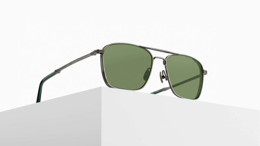 Navigator sunglasses with an antique silver frame and green lenses on a pedestal against a white background