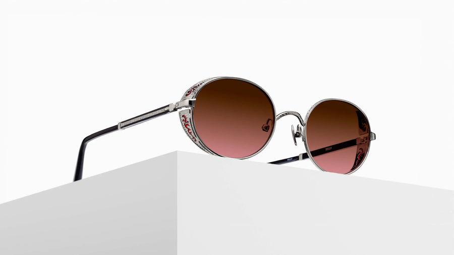 Oval metal sunglasses in a silver tone featuring red accents and black temples, with red gradient lenses, sat atop a grey pedestal