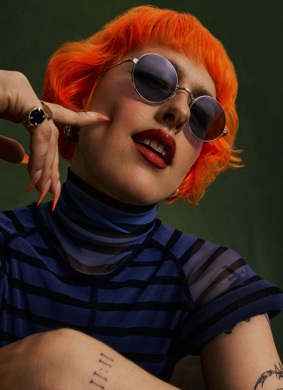 Woman with orange hair and round sunglasses pointing at themselves against a dark background