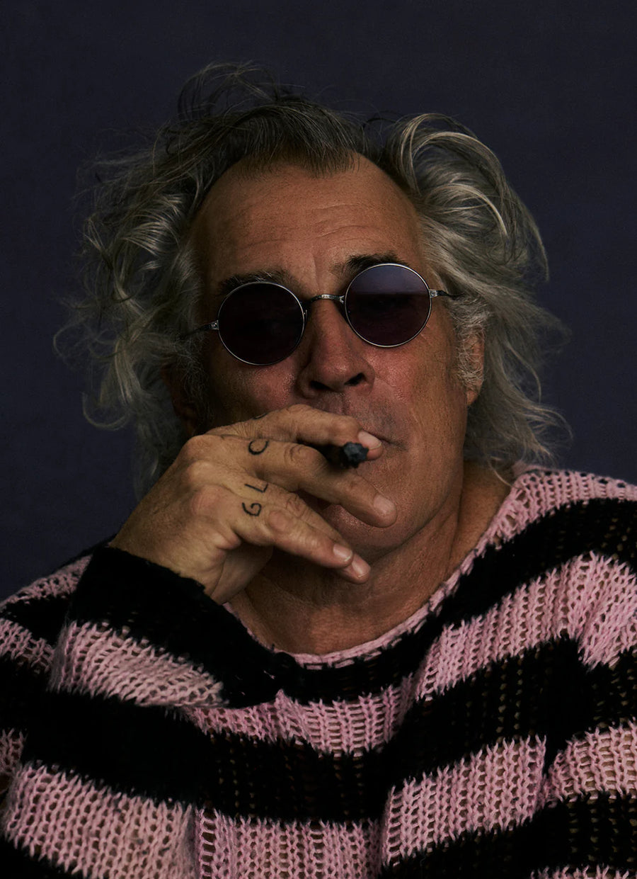Man with gray hair and round sunglasses smoking a cigarette against a dark background.